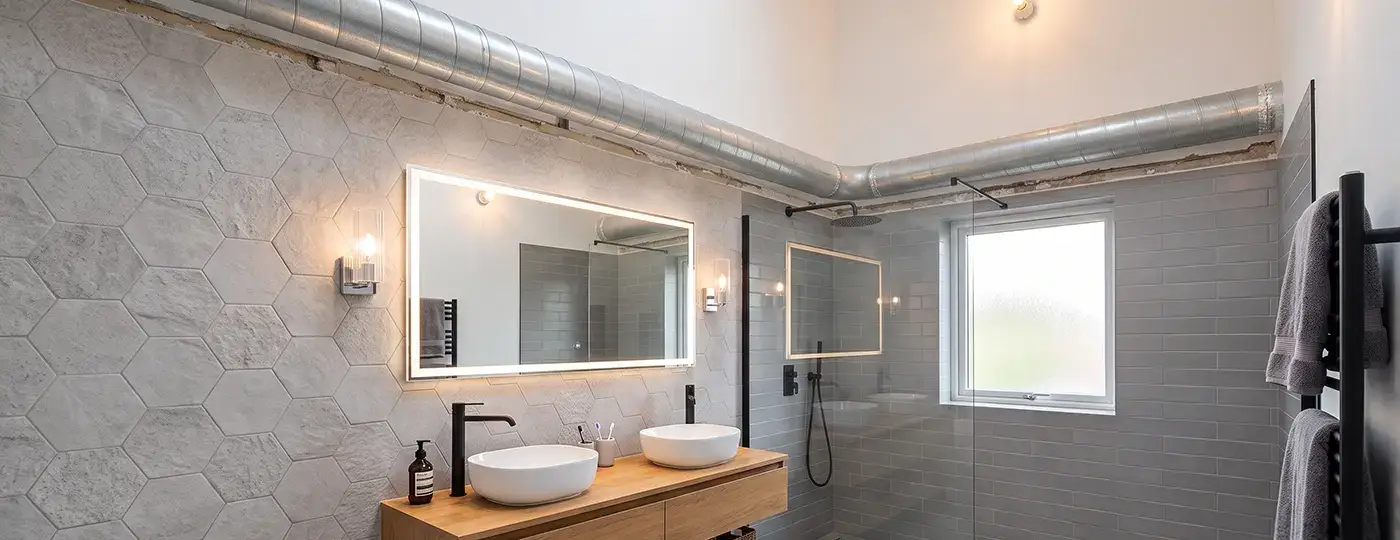 Ein helles Badezimmer, in dem ein metallenes Lüftungsrohr an der Wand sichtbar ist.