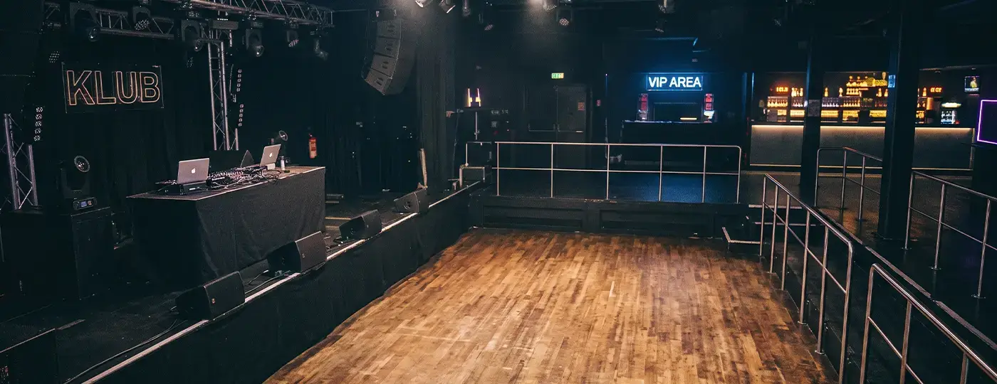 Innenansicht eines leeren Musikclubs mit DJ-Pult, Holztanzfläche und VIP-Bereich.