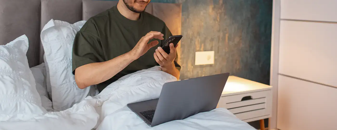 Ein Mann liegt mit einem Laptop im Bett und schaltet das Licht über eine Smartphone-App ein.