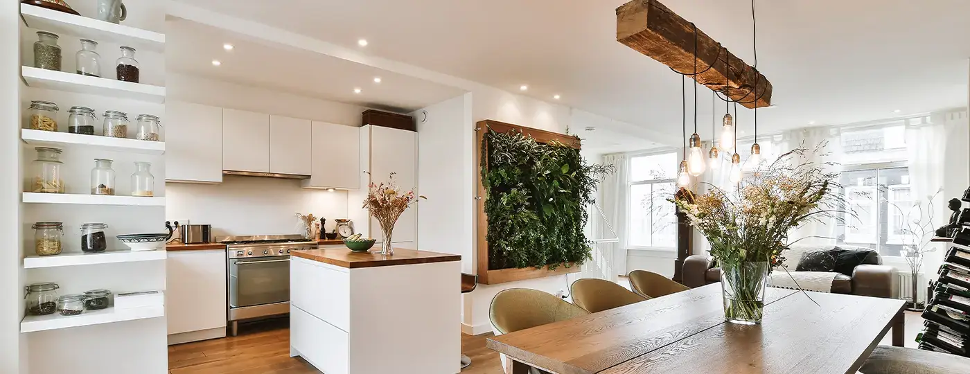 Ein heller, offener Wohnbereich, der eine weiße Küche mit Kochinsel und einen Essbereich mit Holztisch sowie einer rustikalen Pendelleuchte im Balken-Design vereint.