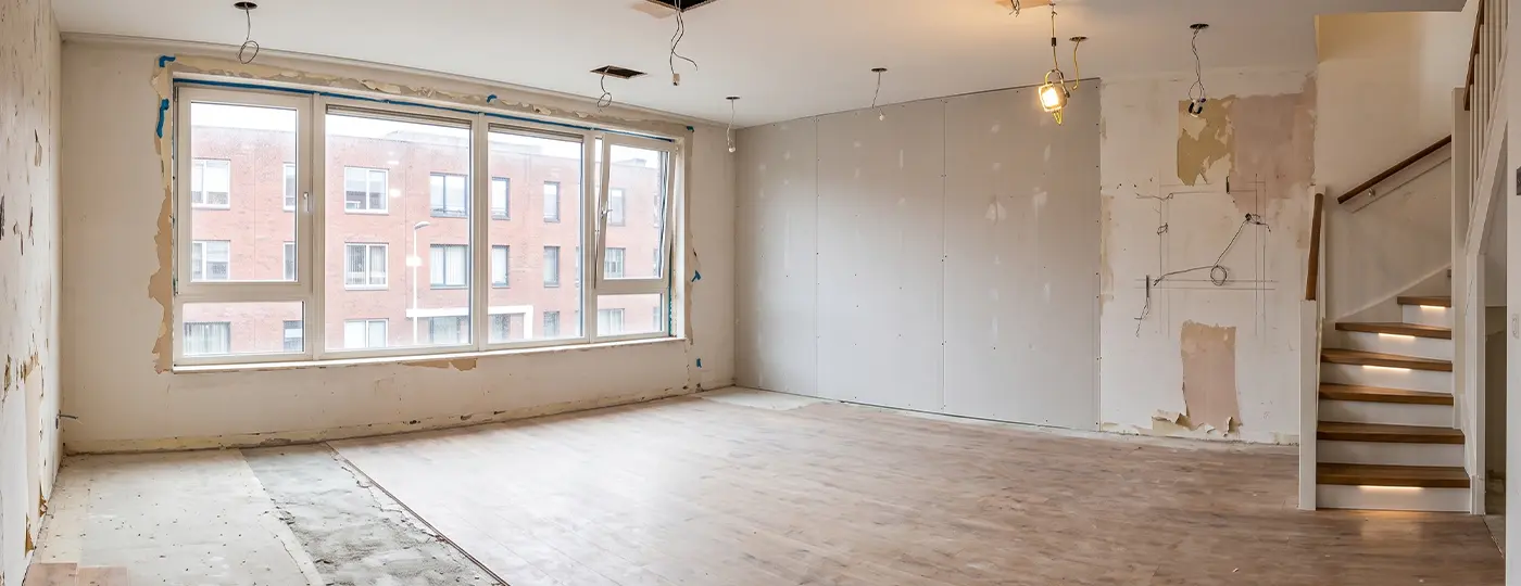 Ein heller, leerer Innenraum einer Maisonette-Wohnung während der Renovierung, mit einem großen Fenster, Trockenbauwänden und beleuchteter Treppe.