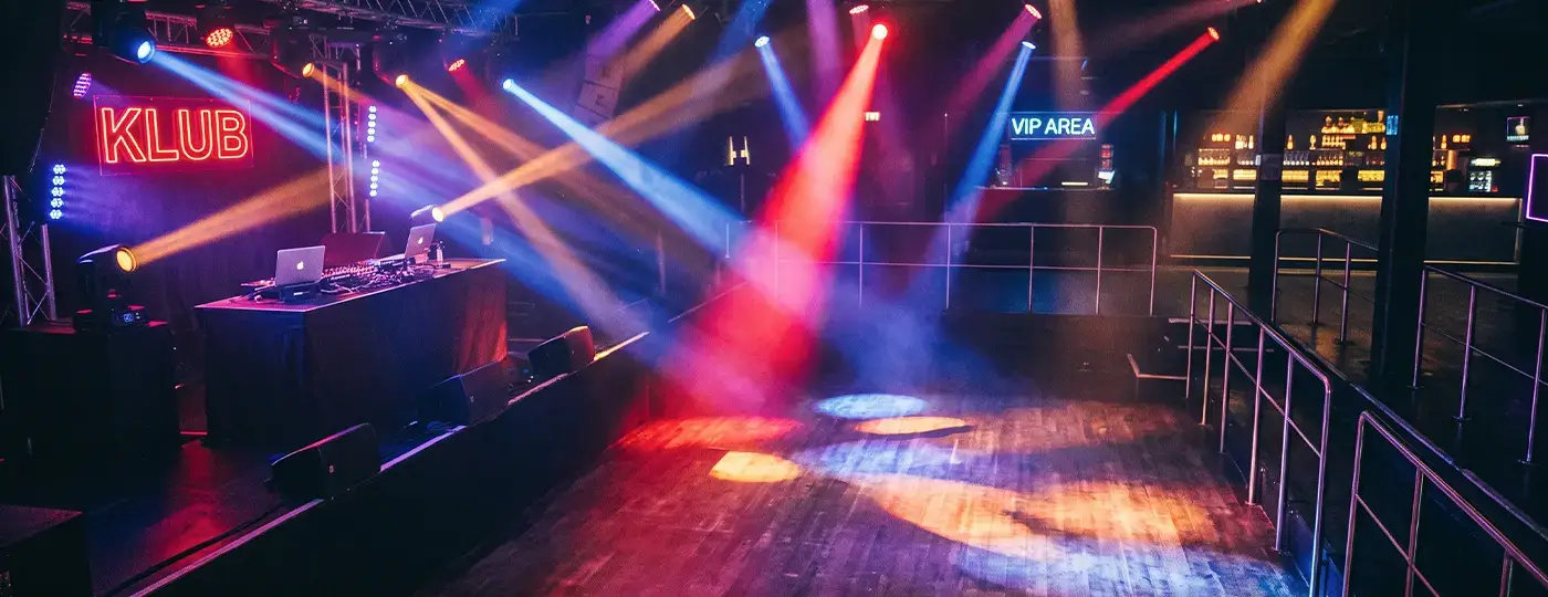 Das Innere eines Musikclubs, beleuchtet von farbigen, sich kreuzenden Lichtstrahlen aus Scheinwerfern, die auf die Tanzfläche und das DJ-Pult gerichtet sind.