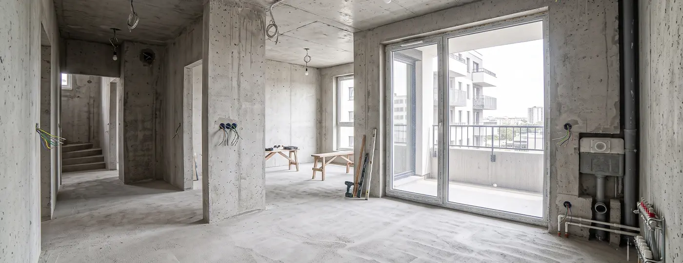 Geräumiges Apartment im Rohbauzustand mit Betonwänden, sichtbarer Elektroinstallation und einem großen Balkonfenster.