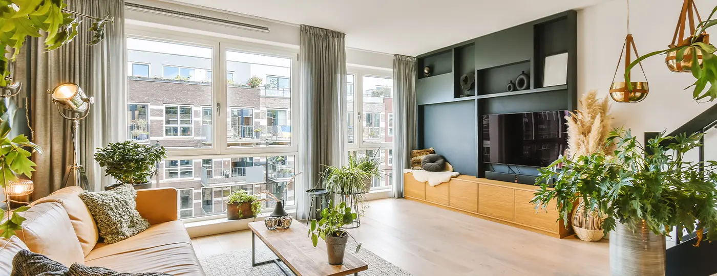 Ein helles, geräumiges Wohnzimmer mit großen Fenstern, einem Ledersofa, zahlreichen Topfpflanzen und einer TV-Wand mit dunklen Einbaumöbeln.