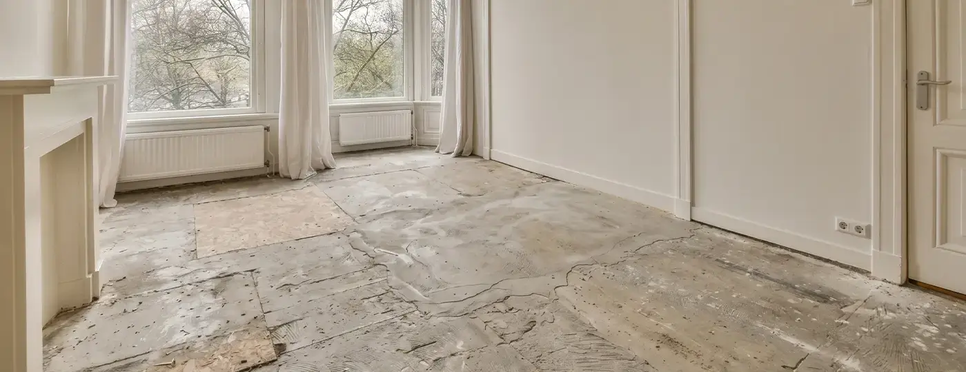 Ein geräumiger, leerer Innenraum im Ausbaustadium mit Rohboden und weißen Wänden; die Fenster sind mit Gardinen sowie einer dekorativen Portalumrahmung versehen.