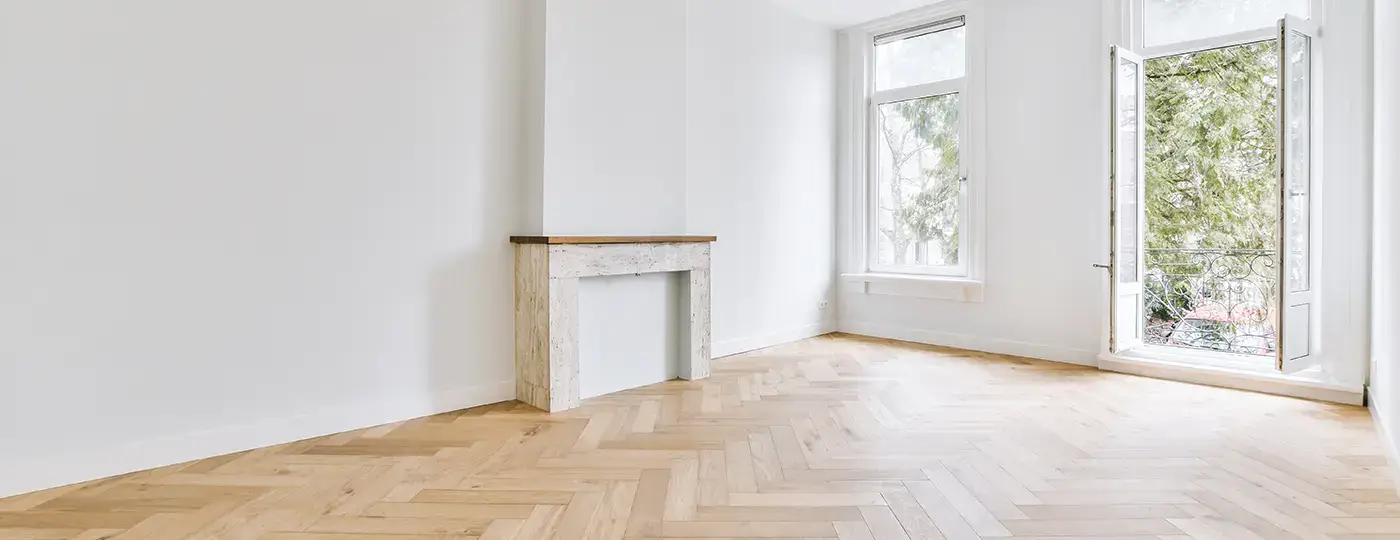 Ein heller, leerer Raum mit weißen Wänden, einem dekorativen Kaminportal, geöffneten Balkontüren und einem neu verlegten Holzboden im Fischgrätmuster.