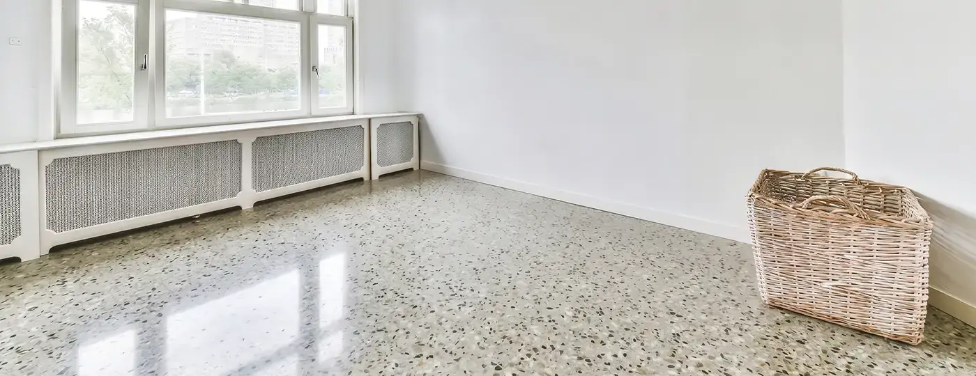 Ein heller, leerer Raum mit einer weißen Wand, einem glänzenden Terrazzoboden, einer großen Fensterfront mit integrierten Heizkörpern sowie einem Weidenkorb in der Ecke.
