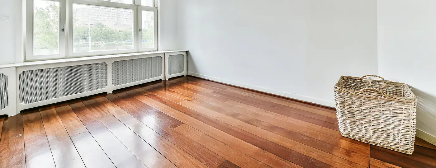 Ein heller, leerer Raum mit weißen Wänden, einem glänzenden Boden aus dunklen Holzdielen, einem großen Fenster mit integriertem Heizkörper sowie einem Weidenkorb in der Ecke.