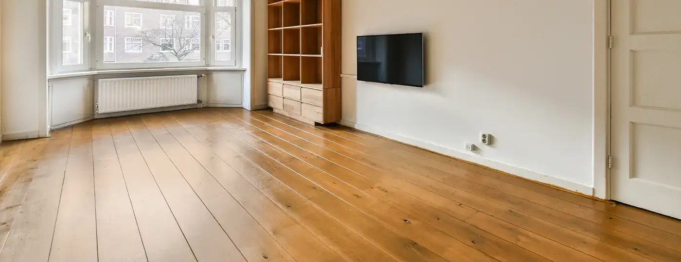 Ein heller, leerer Raum mit Dielenboden, einem Holzregal, einem an der Wand montierten Fernseher sowie einem Erker im hinteren Bereich.
