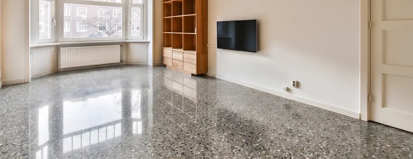 Ein heller, leerer Raum mit glänzendem Terrazzoboden, hölzernen Regalwänden, einem Fernseher an der Wand und einem Erker im hinteren Bereich.