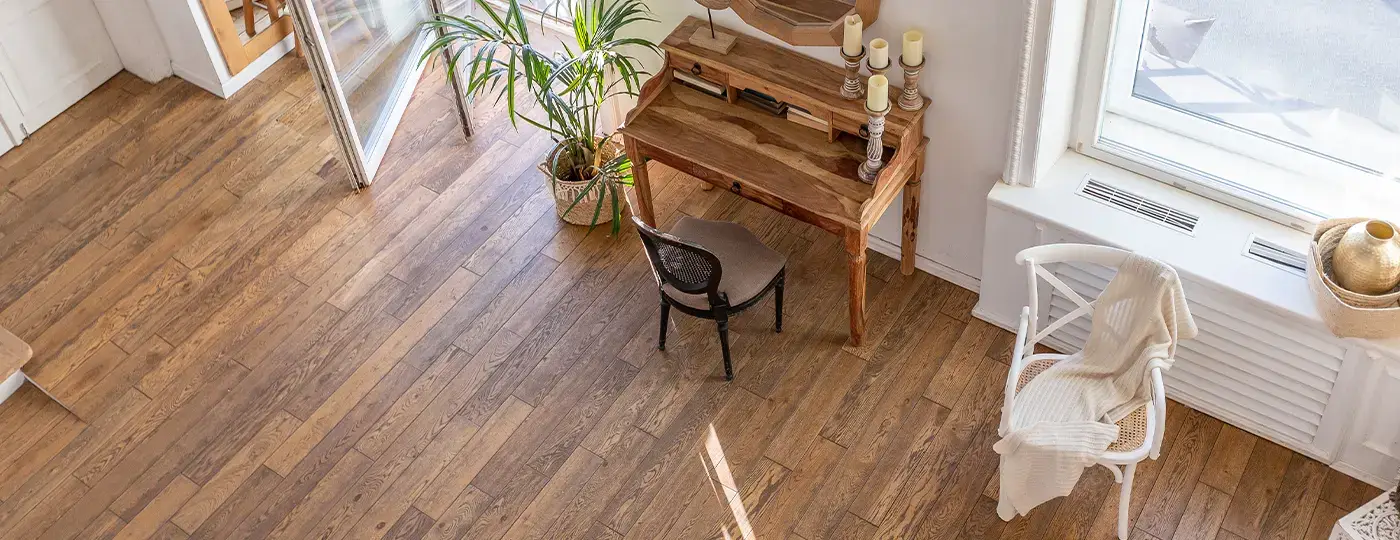 Ein heller Innenraum mit Blick von oben, Holzboden, einem Sekretär aus Holz mit Stuhl sowie einem weißen Sessel am Fenster.