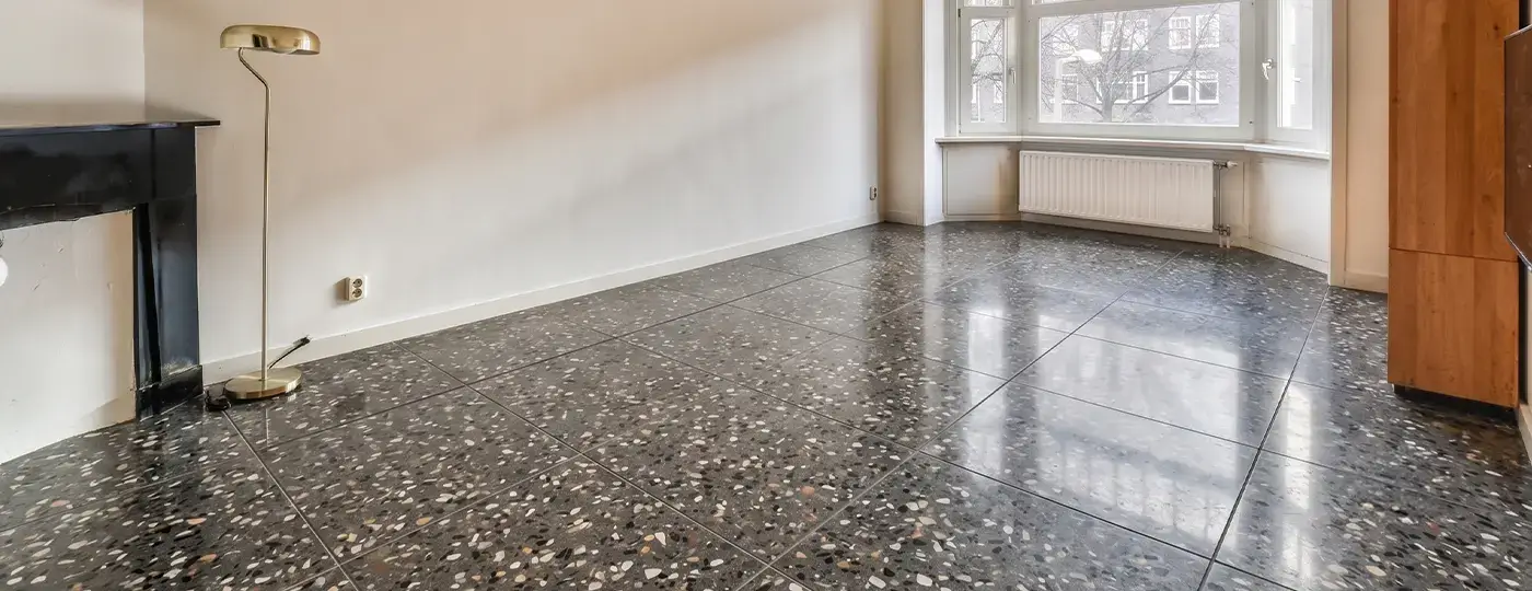 Ein leerer, heller Innenraum mit glänzendem Boden aus dunklem Terrazzo, einem schwarzen Kaminportal, einer Stehlampe sowie einem Fenstererker im Hintergrund.
