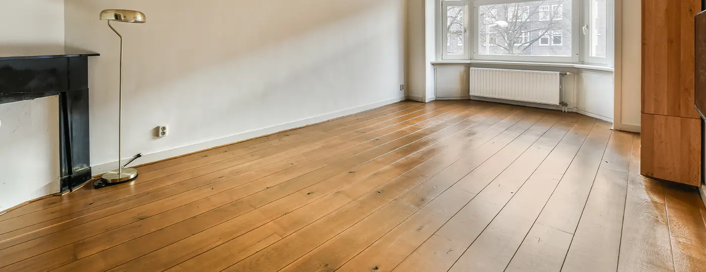 Ein leerer, heller Innenraum mit einem natürlichen Holzboden aus langen Dielen, einem schwarzen Kaminportal, einer Stehlampe sowie einem Erker im Hintergrund.