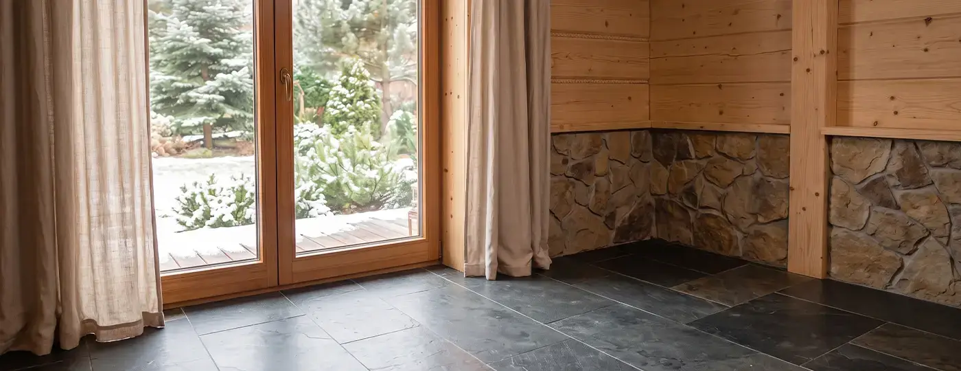 Ein leerer Raum mit dunklem Boden, einer steinernen Sockelwand und einem Fenster zum Wintergarten.