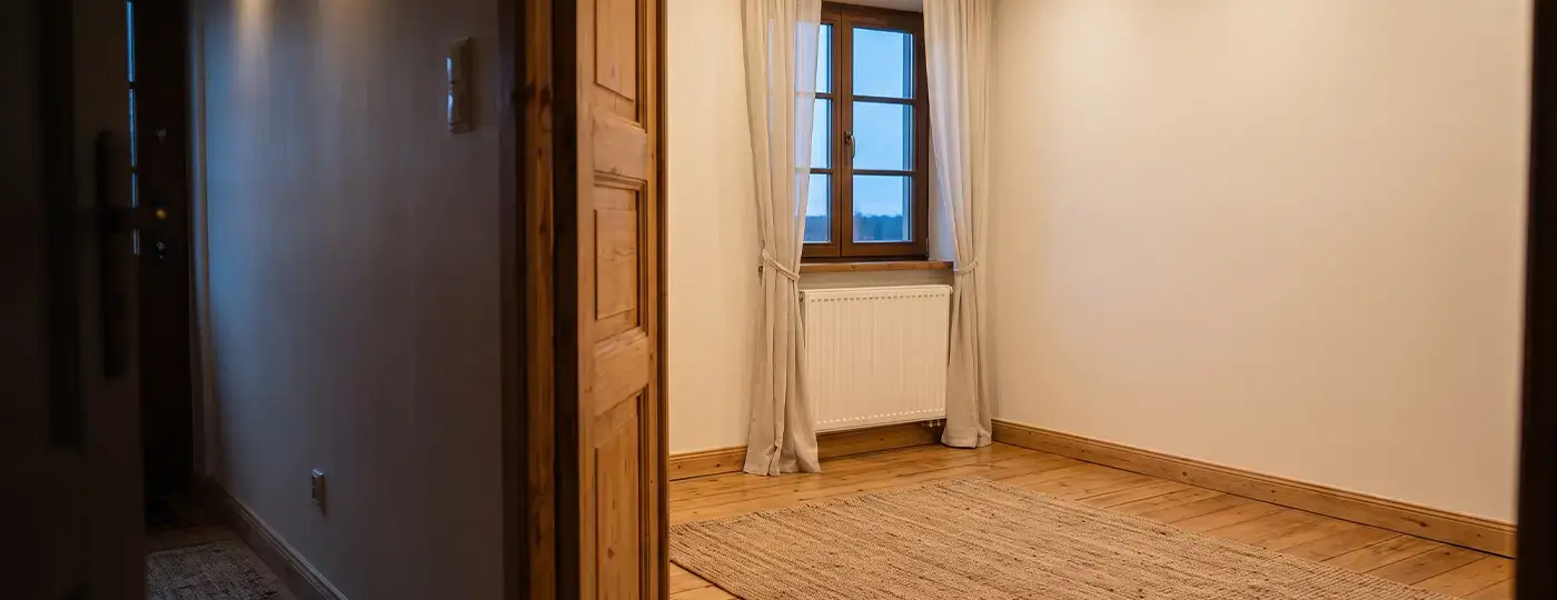 Ein leerer Raum mit Holzboden, geflochtenem Teppich und einem Heizkörper unter dem Fenster.